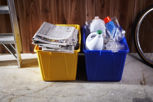 In a residential garage, two large plastic recycling bins are positioned side by side on a concrete floor against a wooden wall. The yellow bin on the left is filled with neatly stacked newspapers and newspapers, with their pages slightly curled and edges uneven, indicating light handling. The blue bin on the right contains clear plastic bottles of various shapes and sizes, some with caps and labels, as well as empty plastic containers and a few disposable water bottles. Both bins are upright, with open tops visible from the front, and appear to be used for household rubbish collection. To the left of the yellow bin, part of a metal shelving unit is visible, and to the right, only a section of a bicycle wheel can be seen. The lighting is indoor, casting a neutral illumination over the scene, typical of a home storage area. This image is relevant to rubbish disposal and recycling services provided by House Clearance Thames Ditton, situated in or near Surrey, emphasizing household waste sorting and collection practices.