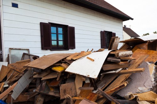 A large pile of mixed construction and demolition debris is situated next to a white residential house with dark brown window shutters, likely in Thames Ditton. The debris includes wooden planks, panels, and particleboards, some with visible textures such as rough, splintered surfaces and smooth painted finishes. There are also some sheets of white and yellow materials among the rubble. The pile is piled haphazardly on a paved driveway or yard, partially covering the ground adjacent to the house’s exterior. In the background, the side of the house features two windows with open shutters, and a neighboring house with a similar roofline is visible further back. The scene appears to be outdoors in daylight, with a neutral, overcast sky, suggesting a recent clearance or renovation activity. This image relates to rubbish removal services provided by House Clearance Thames Ditton, supporting detailed visual understanding of debris typical in house clearance projects in the local area.