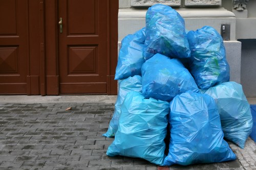 A cluster of multiple large blue plastic rubbish bags filled with domestic waste, stacked on a paved driveway in front of a brown wooden door and garage in a residential area. The bags are made of translucent plastic, revealing some household waste inside, and are tied at the top. The surrounding environment includes a grey brick surface extending across the foreground, with a glimpse of the house's exterior wall featuring decorative mouldings above the door. This scene illustrates rubbish awaiting collection or removal, typical of waste management services in Thames Ditton, where House Clearance Thames Ditton may assist with waste clearance or rubbish disposal for local homes.