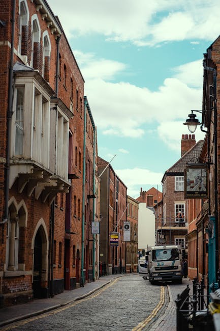 A narrow cobblestone street in an urban setting, flanked by older brick buildings with a mix of architectural styles, including some with ornamental bay windows and decorative brickwork. The buildings