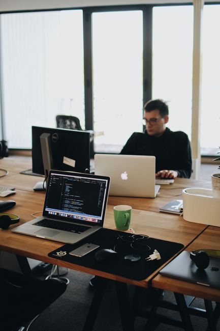 The image depicts a modern office workspace with a large wooden desk positioned near floor-to-ceiling windows, allowing natural light to illuminate the area. On the foreground of the desk, there are t