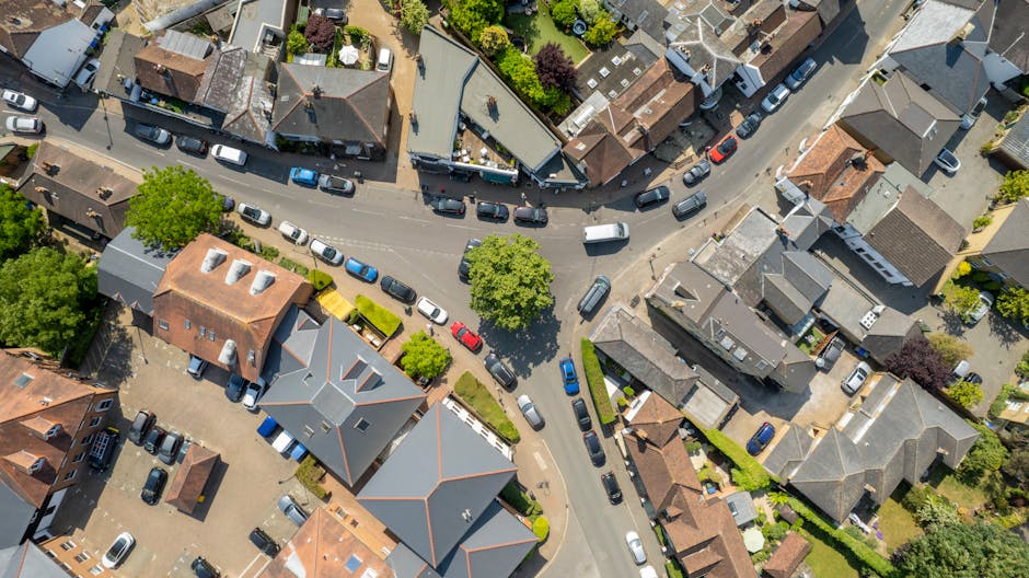 An aerial view of a small urban intersection showing a variety of parked cars, including sedans, hatchbacks, and a van, lining the streets and surrounding residential buildings. Several vehicles are p